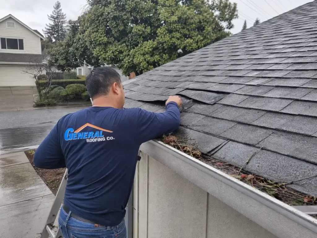 General Roofing technician checking storm damage on a wet residential shingle roof with loose shingles and gutter debris.