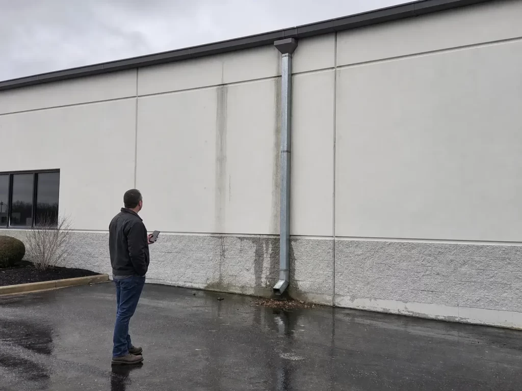 Building owner checking commercial gutter stains and downspout drainage from the ground after rain.
