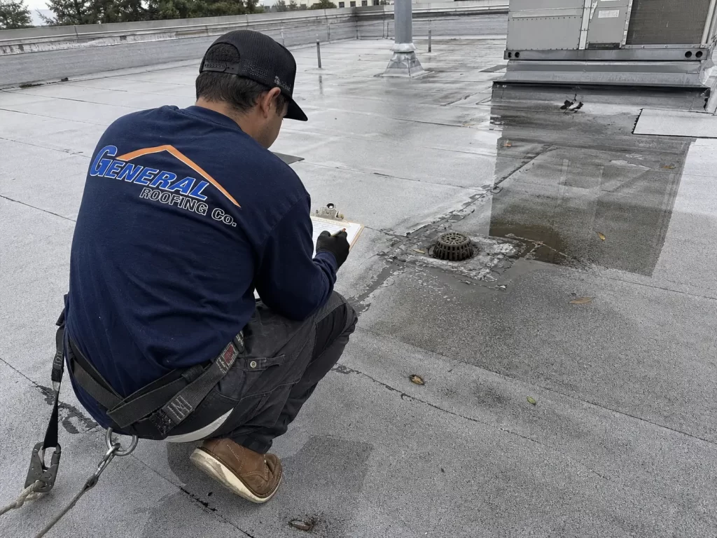 General Roofing contractor inspecting ponding water, membrane seams, and a roof drain on a commercial flat roof.