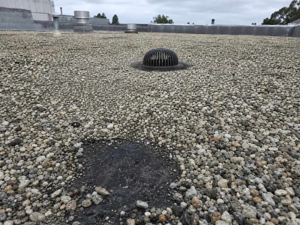 Protective gravel covering a built-up flat roof with exposed asphalt and a clear roof drain.