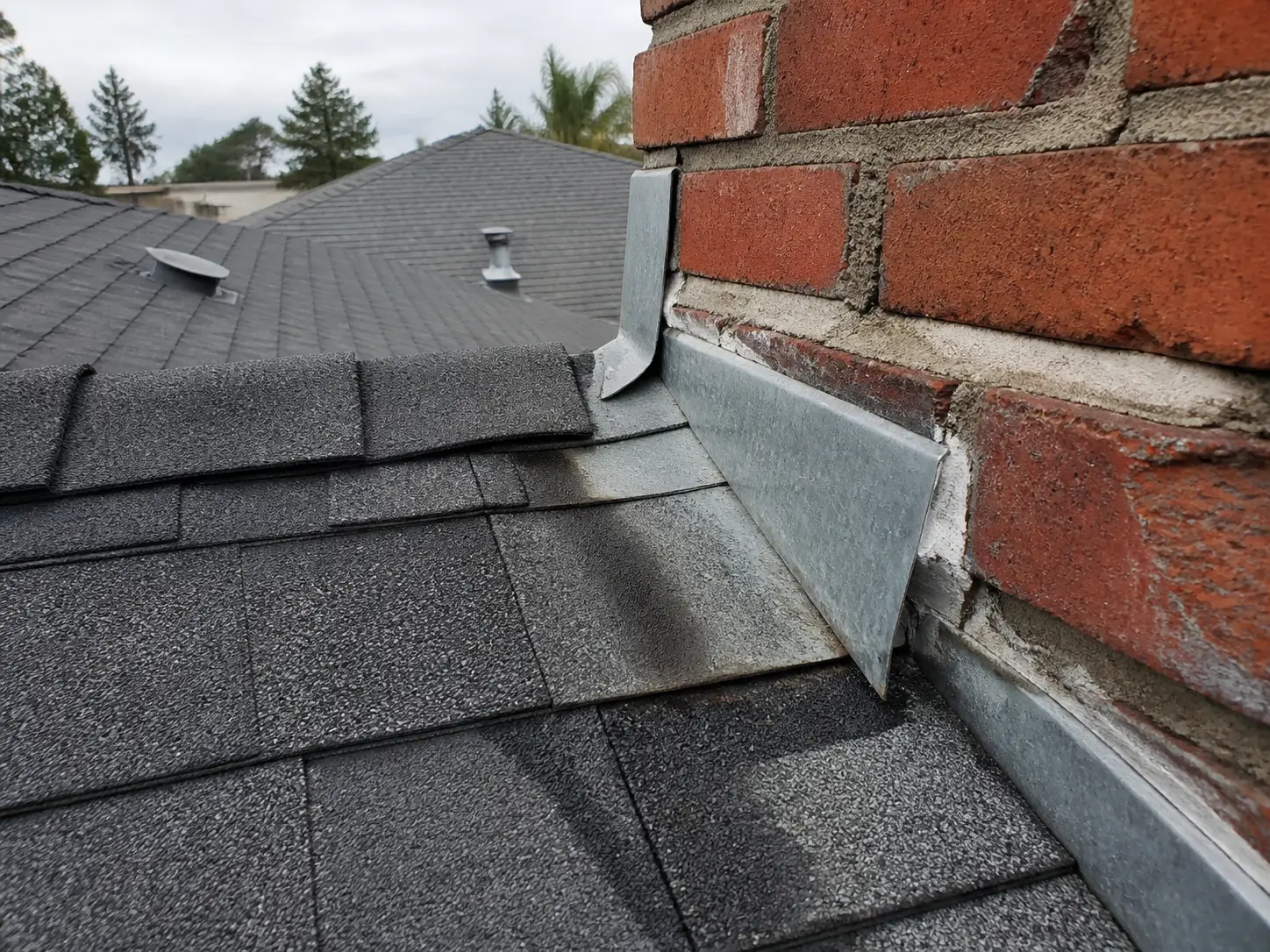 Close-up of loose metal roof flashing where a brick chimney meets an asphalt shingle roof.
